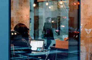 Blick in ein Fenster Frau vor Laptop und Smartphone Spiegelung der Straße und von vorbeilaufenden Menschen symbolisiert zwei Persönlichkeiten: Die digitale Identität und die reale Identität
