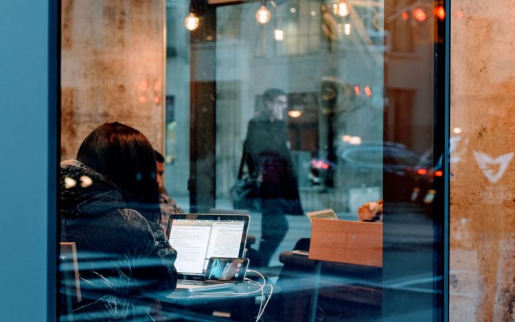 Blick in ein Fenster Frau vor Laptop und Smartphone Spiegelung der Straße und von vorbeilaufenden Menschen symbolisiert zwei Persönlichkeiten: Die digitale Identität und die reale Identität