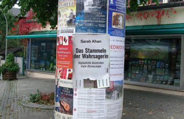 Litfaßsäule mit Plakaten und Anzeigen. In der Mitte das Cover des Buches Das Stammeln der Wahrsagerin über eBay-Geschichten